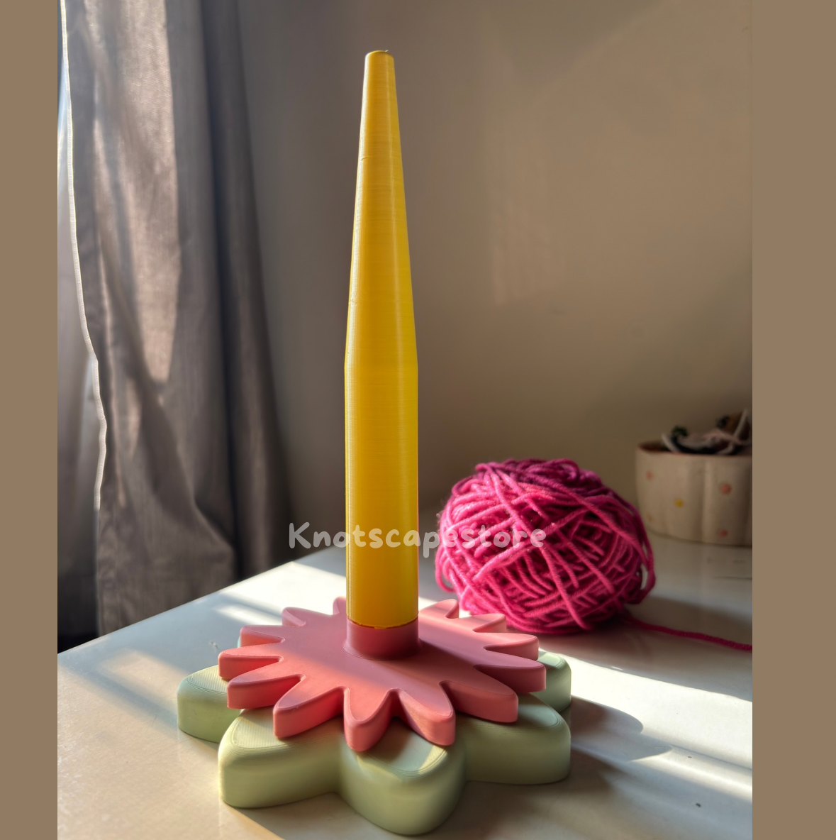 floral yarn spinner in pastel green and pink with a yellow center rod. Ideal for crochet and knitting organization.Pink yarn ball in background .Features the Knotscapestore watermark.