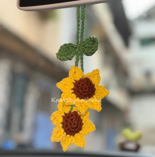 Crocheted sunflower decorations hanging from a car mirror with 'KnutsbyAnshuka' branding.