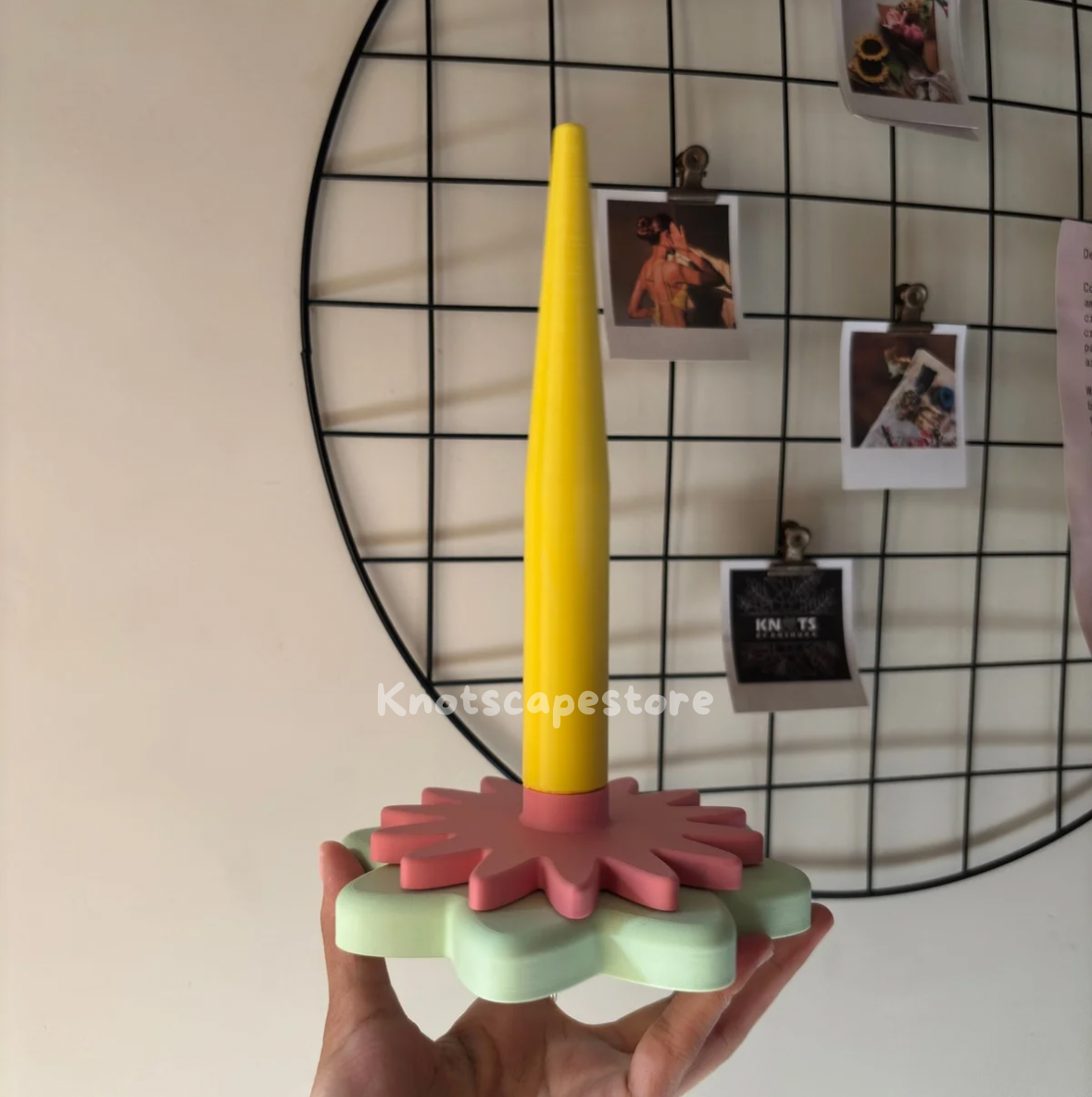 A hand holding a floral-themed yarn spinner with a tall yellow spindle. The base consists of two stacked, matte-finished flower shapes in soft sage green and dusty rose. The "Knotscapestore" watermark is visible in the center.