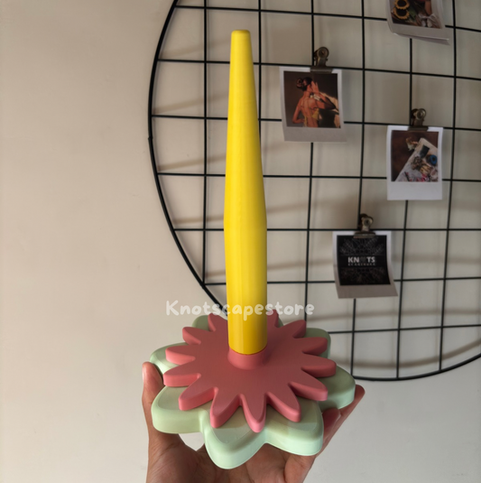 A hand holding a floral-themed yarn spinner with a tall yellow spindle. The base consists of two stacked, matte-finished flower shapes in soft sage green and dusty rose.